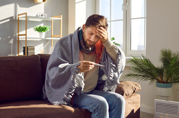 Sick man covered with blanket measuring temperature with thermometer. Unhappy man suffering from...