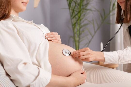 Unrecognizable pregnant female with bare belly and stethoscope doctor listening to baby heartbeat woman wearing white shirt appointment in hospital.