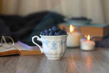 Cup of blueberries, set of books, phone, eyeglasses, lit candles and blanket on the table. Hygge at home. Selective focus.