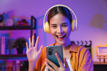 Cheerful beautiful Asian woman wearing headphones and look to smartphone at video calling meeting...