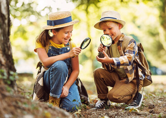 Two little kids with backpacks examining fir cone through magnifying glass in forest