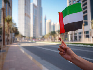 Close up of a female hand holding tiny flag of UAE against Dubai skyline. Photo of UAE national day celebration spirit.