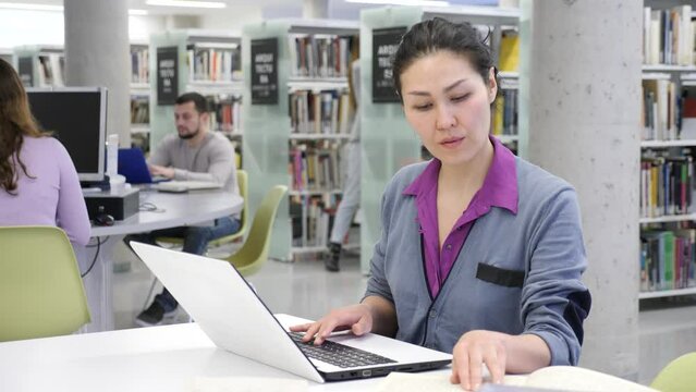 Young focused woman working with laptop and books, finding information at library and making notes