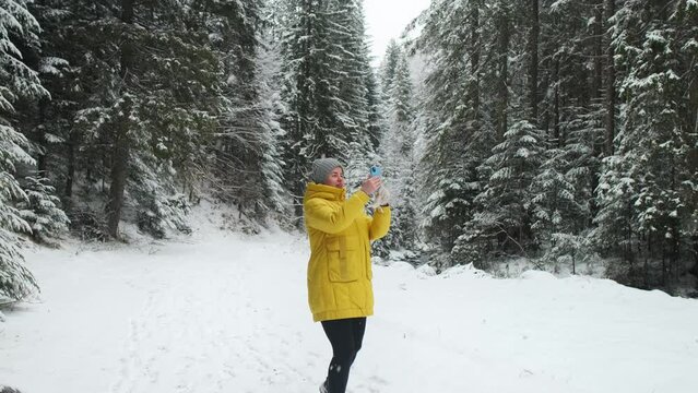 Woman in bright yellow winter jacket hikes in the snowy mountain forest and makes photos
