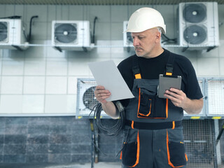 Man electrician. Worker near wall with air conditioners. Air conditioner repairman. Electrician in helmet and work overalls. Engineer controls operation of conditioners. Man with wire and tablet