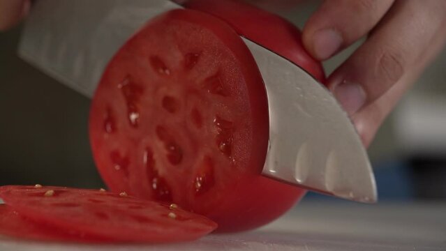 Close up shot of chef's hands cutting a fresh tomato with knife. Cooker preparing vegetarian salad in kitchen