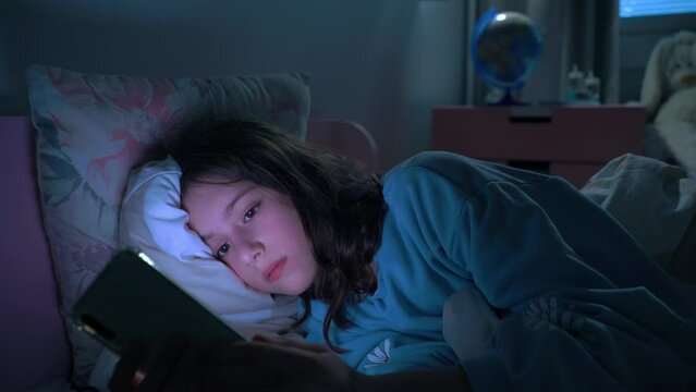 Teen girl lying in bed at night using smartphone, scrolling social media and texting. Not healthy sleep. Insomnia and the harm of the smartphone before going to bed. Dependence on social networks