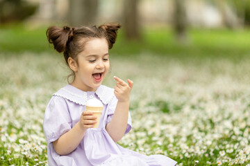 Funny happy toddler girl wearing cotton summer dress sitting on a green lawn eating vanilla and chocolate ice cream cone in a sunny garden or park. Space for text