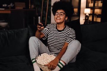 Joyful african woman watching tv and eating popcorn while sitting on couch