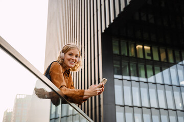 Woman listening music with headphones and mobile phone while standing outdoors near modern office building