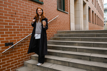 Young smiling woman in headphones drinking coffee while standing outdoors on stairs