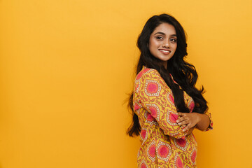 Young indian woman smiling while standing with folded arms isolated over yellow background