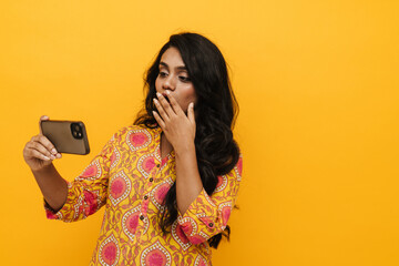 Suprised indian woman covering her mouth while looking at mobile phone screen isolated over yellow background