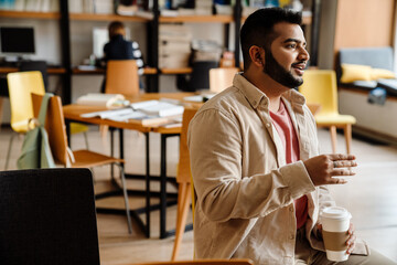Indian man drinking coffee and talking to somebody while sitting in library