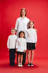Fototapeta premium Portrait of beautiful young girl posing with little kids, boy and girls against red studio background. Same style clothes. Concept of family, motherhood, childhood, fashion, lifestyle, love