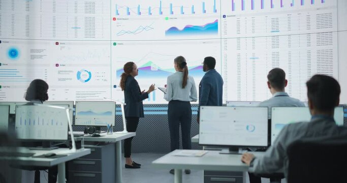 Diverse Team Of Data Scientists Analyzing Information And Graphs On Big Digital Screen In Modern Monitoring Office. Multiethnic Employees Working Infront Of Desktop Computers In Consulting Firm.