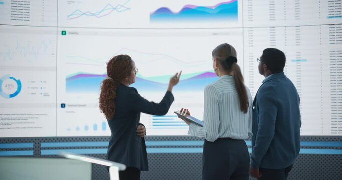 Diverse Team Of Data Scientists Analyzing Information And Graphs On Big Led Wall In Modern Monitoring Office. Multiethnic Colleagues Discussing Financial Strategies, Risk Factors, Potential Deals.