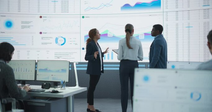Diverse Team Of Financial Data Analysts Studying Information And Graphs On Big Digital Screen In Modern Monitoring Office. Multiethnic Employees Working Infront Of Desktop Computers In Consulting Firm