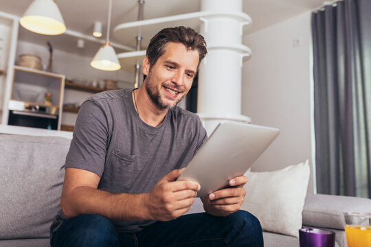 Handsome man is using a digital tablet and smiling while resting on couch at home