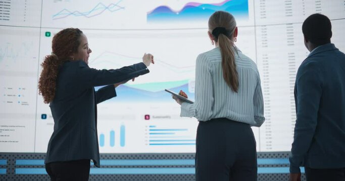 Low Angle Shot: Diverse Team Of Data Scientists Analyzing Information And Graphs On Big Led Wall In Modern Monitoring Office. Multiethnic Colleagues Discussing Financial Strategies and Risk Factors.