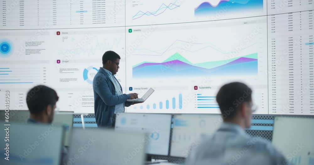 Poster Male Financial Data Analyst Using Laptop And Studying Information And Graphs On Big Digital Screen In Monitoring Office. Black Man Working For Big Consulting Firm, Diverse Employees Behind Computers.