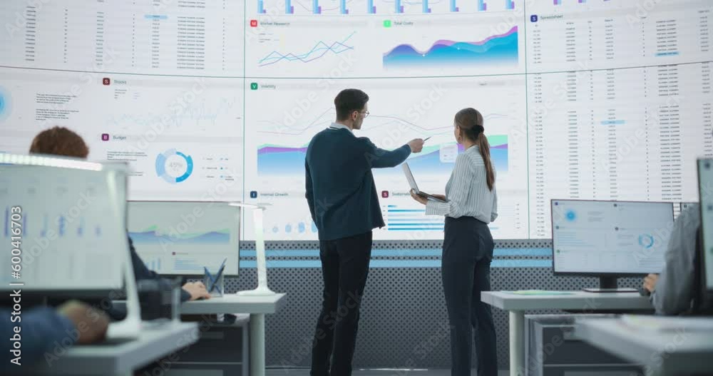 Leinwandbilder Back View Following Shot Of Male And Female Analysts Discussing Data On Big Digital Screen In Monitoring Office. Diverse Employees Working Behind Desktop Computers In Artificial Intelligence Company.