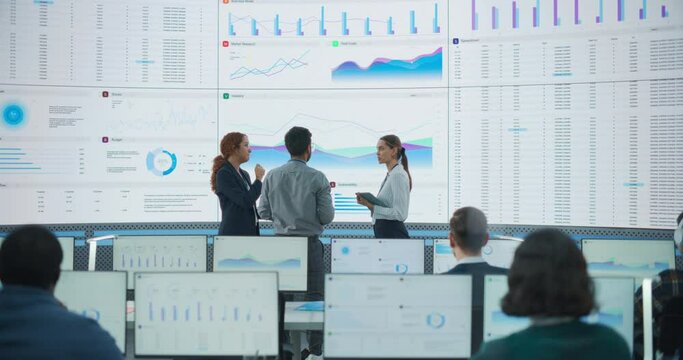 Wide Back Shot of Diverse Team of Data Scientists Discussing Graphs And Reports On Big Digital Screen In Monitoring Office. Multiethnic Employees Working On Desktop Computers For Consulting Company.