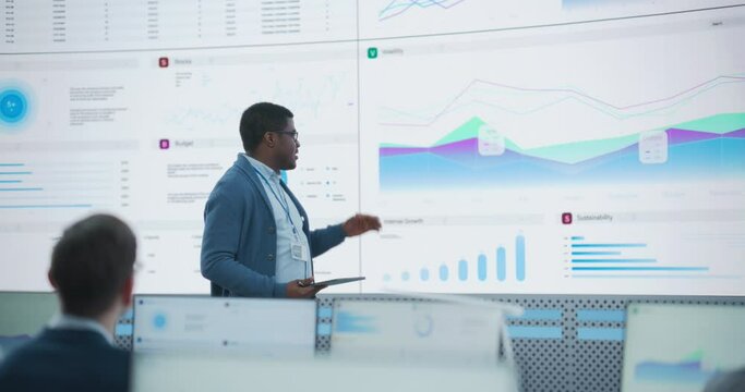 Black Male Financial Analyst Giving Presentation To Venture Capital Company Employees On Big Digital Screen In Monitoring Room. Diverse Colleagues Listening to New Objectives Behind Desktop Computers.