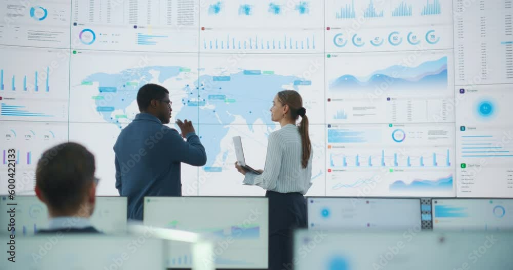 Poster Back View Of Black Male Data Scientist Talking To Caucasian Female Supervisor In Front Of Digital Screen With World Map In Monitoring Office. Employees Working Behind Computers in Distribution Company