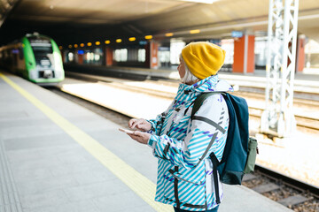 Old happy woman with backpack in yellow hat waiting train on station platform and using smart phone on urban background. Railroad transport concept