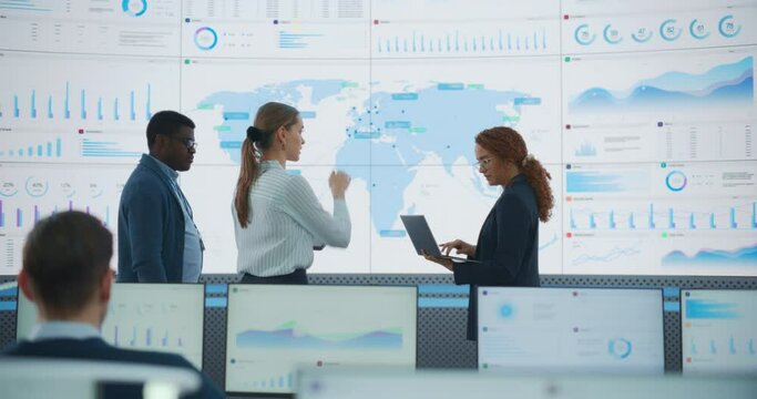 Diverse Team Of Data Science Department Discussing New Markets In Front of Big Digital Screen With World Map In Monitoring Office. Multiethnic Employees Working Behind Computers For Consulting Company