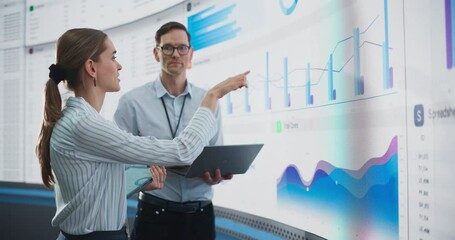 Caucasian Male And Female Data Scientists With Laptop And Tablet Standing Next To Big Digital Screen With Graphs And Charts In Monitoring Office. Colleagues Discussing Business Opportunities, Risks. - Powered by Adobe