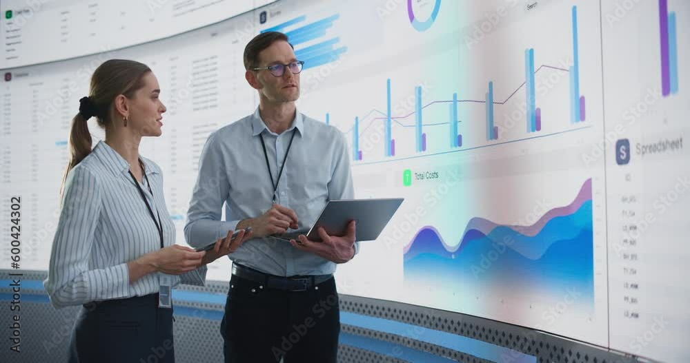 Poster caucasian male and female data scientists with laptop and tablet standing next to big digital screen