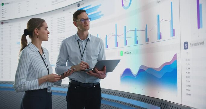 Caucasian Male And Female Data Scientists With Laptop And Tablet Standing Next To Big Digital Screen With Graphs And Charts In Monitoring Office. Colleagues Discussing Business Opportunities Or Risks.