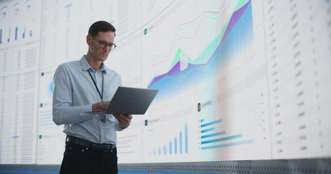In The Office: Male Monitoring Specialist Holding Laptop Computer And Standing Next To Big Digital Screen With Graphs And Charts. Caucasian Man Analyzing Data, Performance Records, And Filing Report. - Powered by Adobe