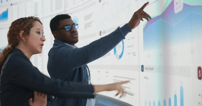 In the Monitoring Office: Black Man And Hispanic Woman From Data Analysis Department Standing Next To Big Digital Screen With Graphs And Charts. Male And Female Specialists Discussing Strategies. - Powered by Adobe
