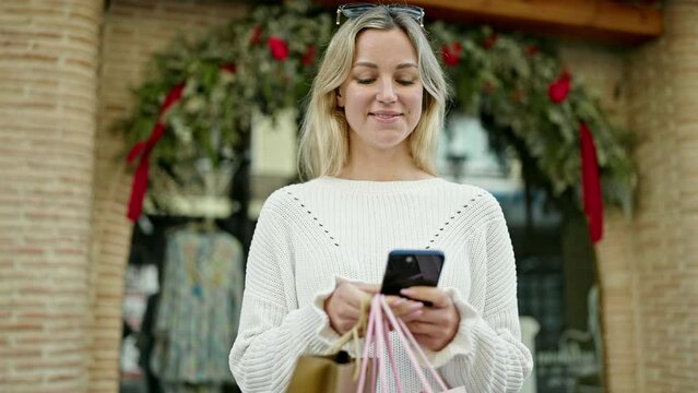 Young blonde woman going shopping holding bags using smartphone at street