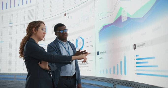 Black Man And Hispanic Woman Working On Business Strategy and Standing Infront of Big Digital Screen With Data. Male And Female Colleagues Discussing Plan For Profit Margins Enlargement In The Office.