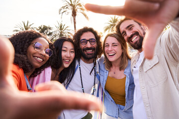 Happy selfie of an internacional group of friends making frame gesture with fingers during their beach holidays outdoors. People looking at camera smiling. - obrazy, fototapety, plakaty