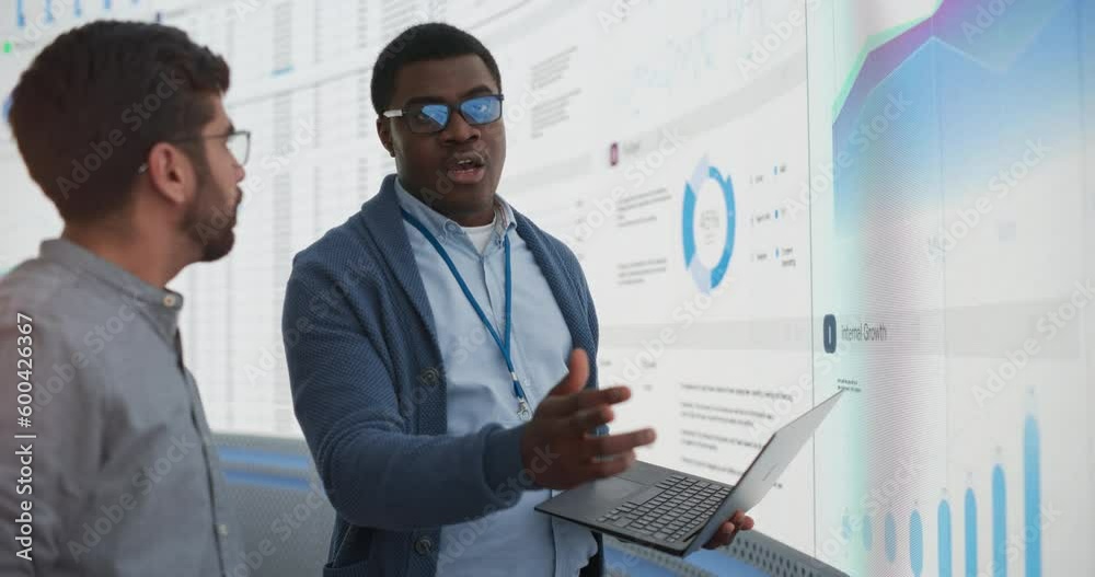 Aufkleber African Project Manager With Laptop Computer Discussing With Indian Analyst Growth Opportunities In Front Of Big Digital Wall In The Office. Two Successful Men Planning New Strategies For Business.