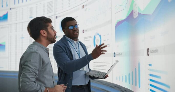 African Data Science Team Lead With Laptop Computer Reporting To Indian CEO About Growth And Sales In Front Of Big Digital Wall In The Office. Two Successful Men Discussing Performance Of The Company.