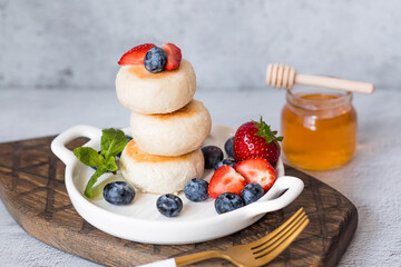 Cottage cheese pancakes, cheesecakes with fresh blueberries, strawberries, honey and mint leaves on a stylish wooden board on a gray concrete table. Natural products. Healthy and delicious breakfast.