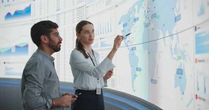 Indian Male Data Analyst And Caucasian Female Project Manager Talking In Front Of Big Digital Screen With Graphs, Charts And Map In Monitoring Office. Diverse Colleagues Discussing Profit Margins.