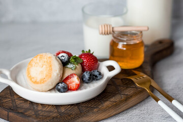 Cottage cheese pancakes, cheesecakes with fresh blueberries, strawberries, honey and mint leaves on a stylish wooden board on a gray concrete table. Natural products. Healthy and delicious breakfast.