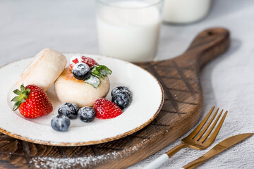 Cottage cheese pancakes, Russian cheesecakes with fresh blueberries, strawberries and mint on a stylish wooden board on a gray concrete table. Natural products. Healthy and delicious breakfast.