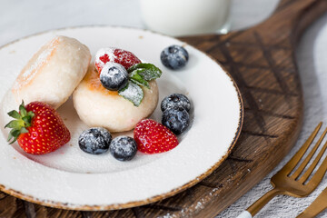 Cottage cheese pancakes, Russian cheesecakes with fresh blueberries, strawberries and mint on a stylish wooden board on a gray concrete table. Natural products. Healthy and delicious breakfast.