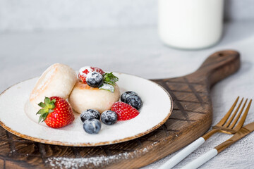 Cottage cheese pancakes, Russian cheesecakes with fresh blueberries, strawberries and mint on a stylish wooden board on a gray concrete table. Natural products. Healthy and delicious breakfast.