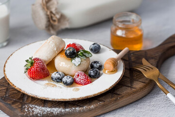 Cottage cheese pancakes,cheesecakes with fresh blueberries, strawberries, honey and mint leaves on a stylish wooden board on a gray concrete table. Natural products. Healthy and delicious breakfast.