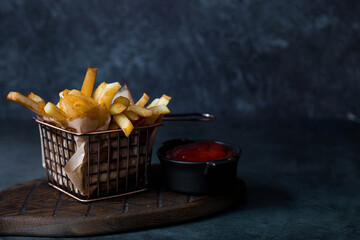 French fries in a metal basket with salt and ketchup on a stylish board on a dark background. Fried...