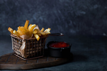 French fries in a metal basket with salt and ketchup on a stylish board on a dark background. Fried...